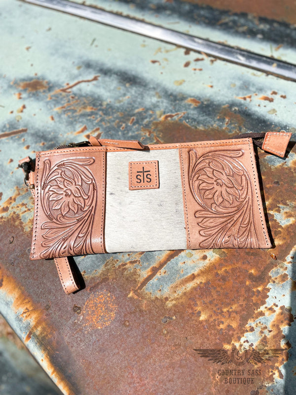 Image of the front side of option A of yipee kiyay clutch wallet with veggie tan leather that is stamped with a floral design on each side with white cowhide with faint black speckles in the middle with a leather patch with STS branded into it.  